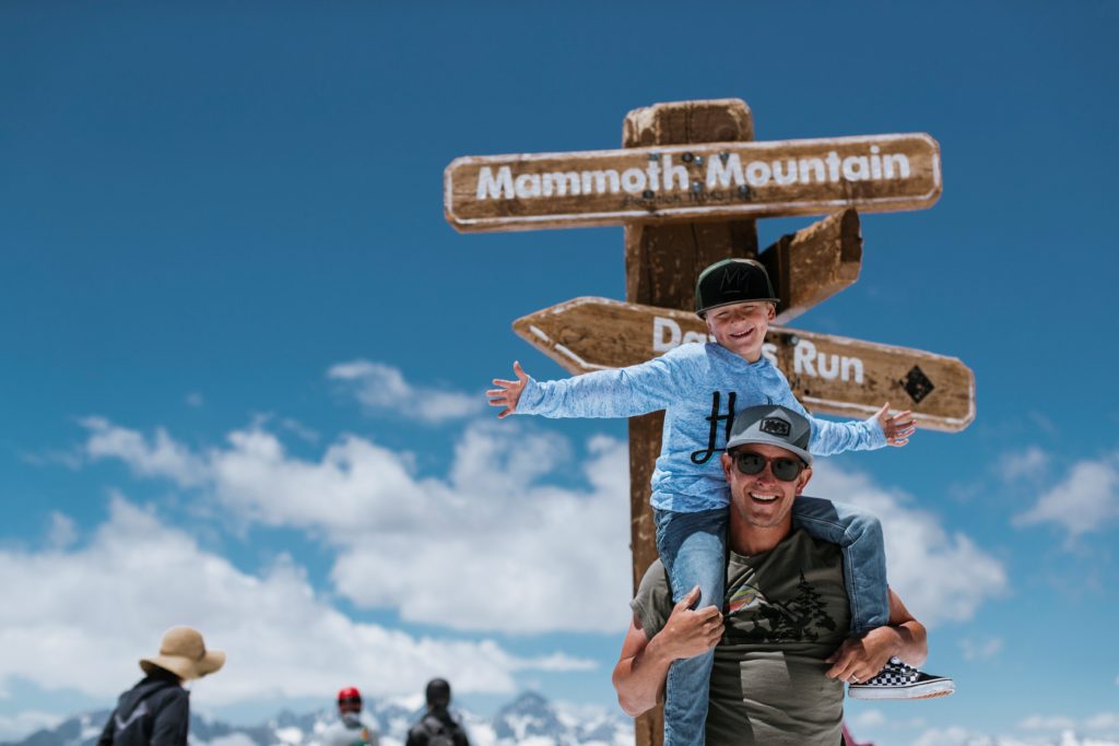 Man and son smiling in front of Mammoth Mountain's mountaintop trail sign.